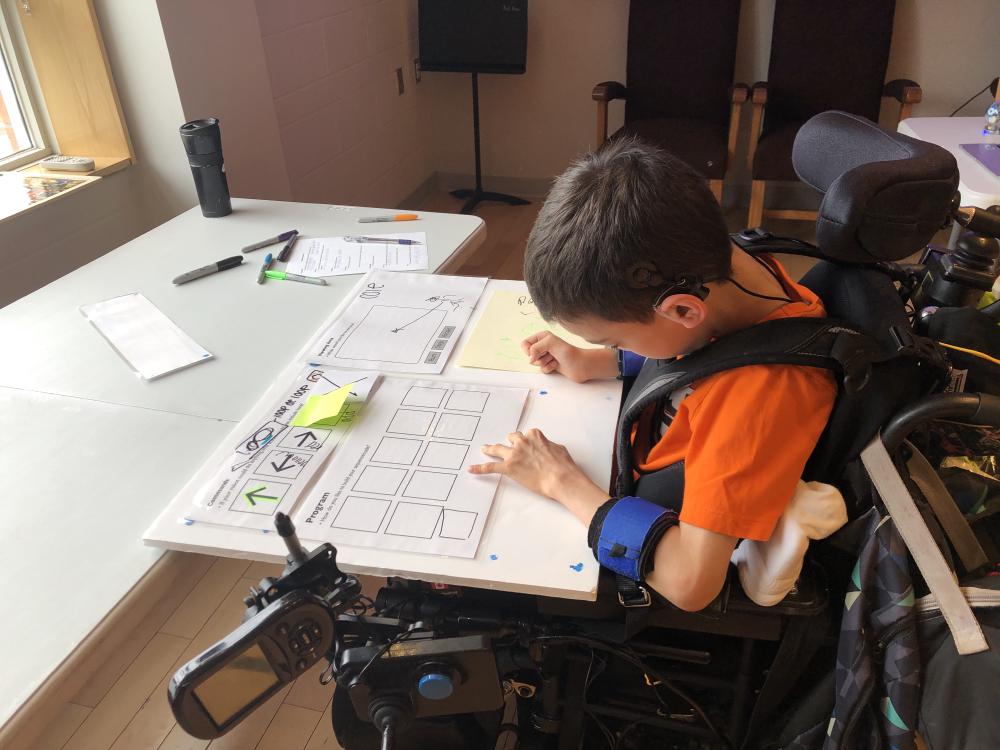 The planned drawing activity on a foam board was inaccessible for the student. child in a mobility device with a foam board and sticky notes